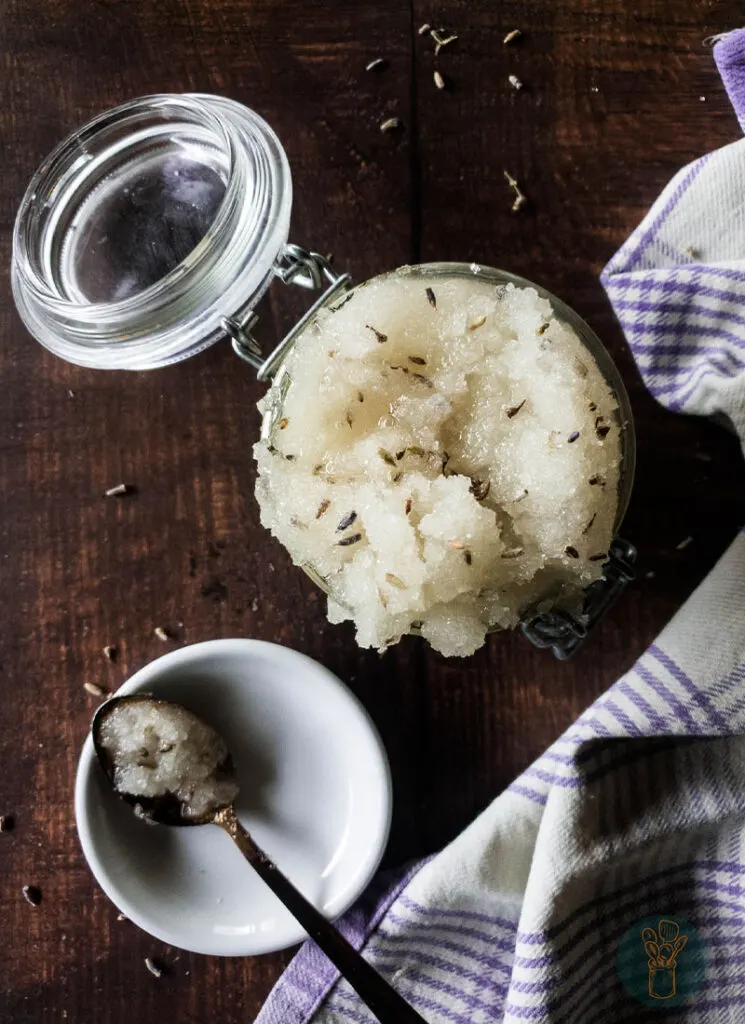 A jar of lavender sugar scrub next to a spoonful of the same scrub in a white saucer.