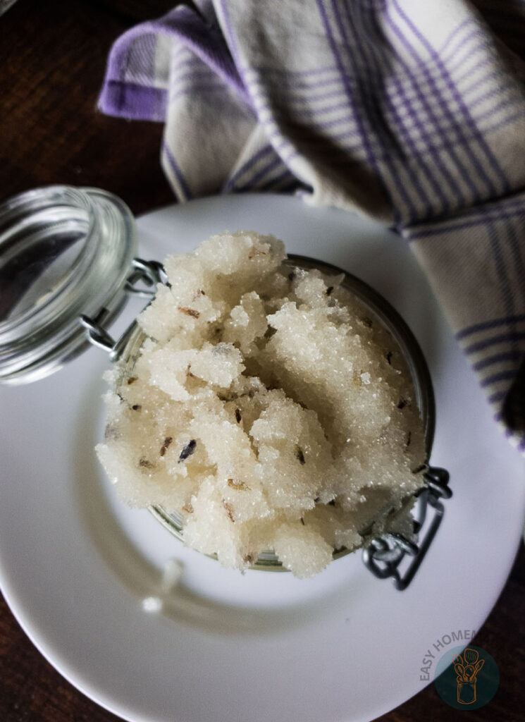 An open jar of lavender sugar scrub on a white plate next to a purple and white checkered napkin.