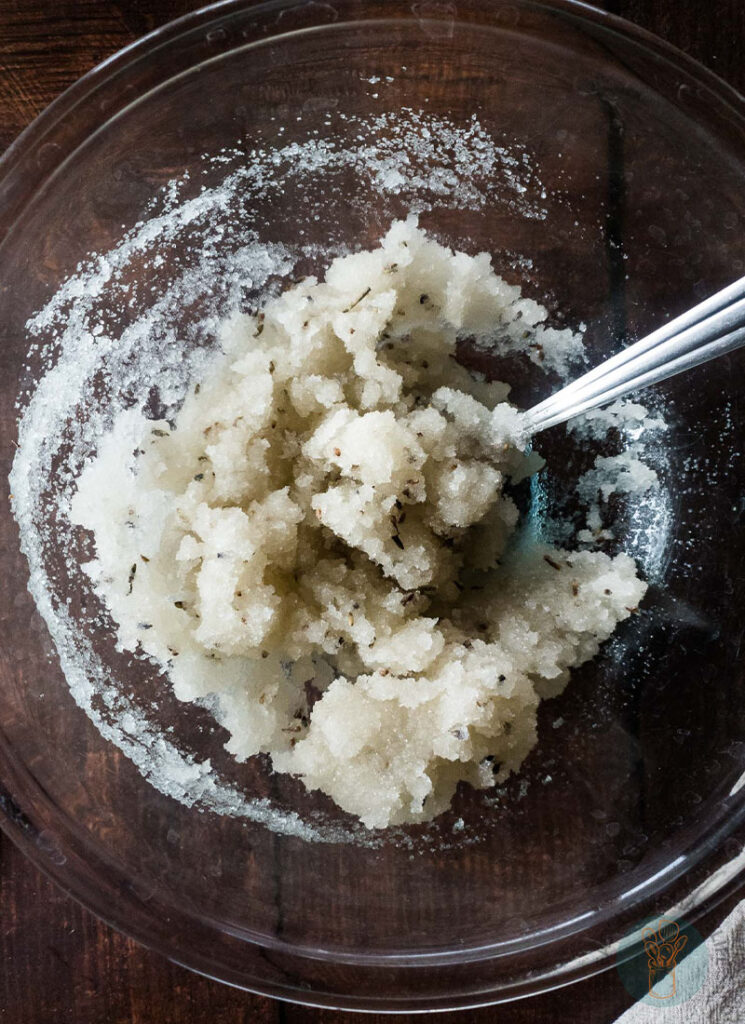 Lavender sugar scrub ingredients being mixed in a glass bowl using a spoon.