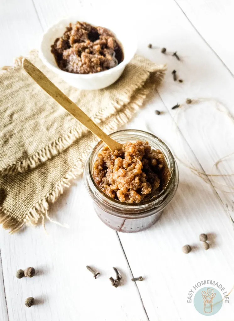 A jar of pumpkin pie spice facial scrub with a wooden spoon next to a white bowl on a burlap with the same facial scrub.