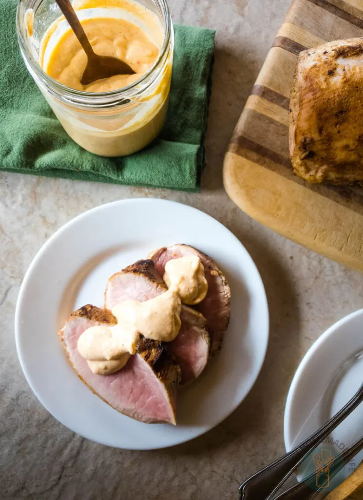 Tiger sauce over slices of cooked meat on a white plate next to a jar of the same sauce and a slab on meat on a wooden cutting board.