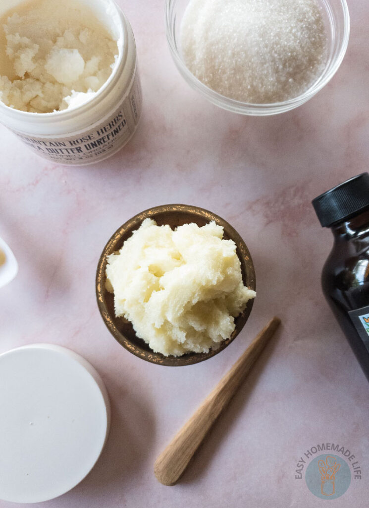 Whipped sugar scrub in wooden bowl next to a bowl of sugar, a bottle of vanilla extract and a jar of shea butter.