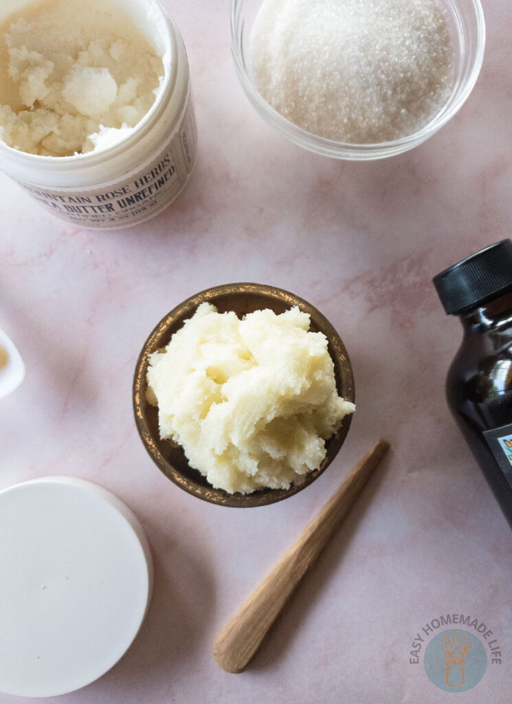 Whipped sugar scrub in wooden bowl next to a bowl of sugar, a bottle of vanilla extract and shea butter.