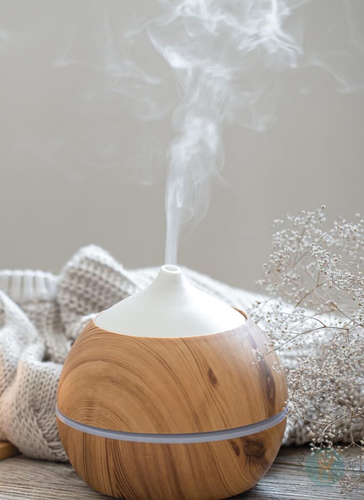 Essential oil diffuser diffusing mist next to a branch of tiny dried white flowers and a white knitted cloth.