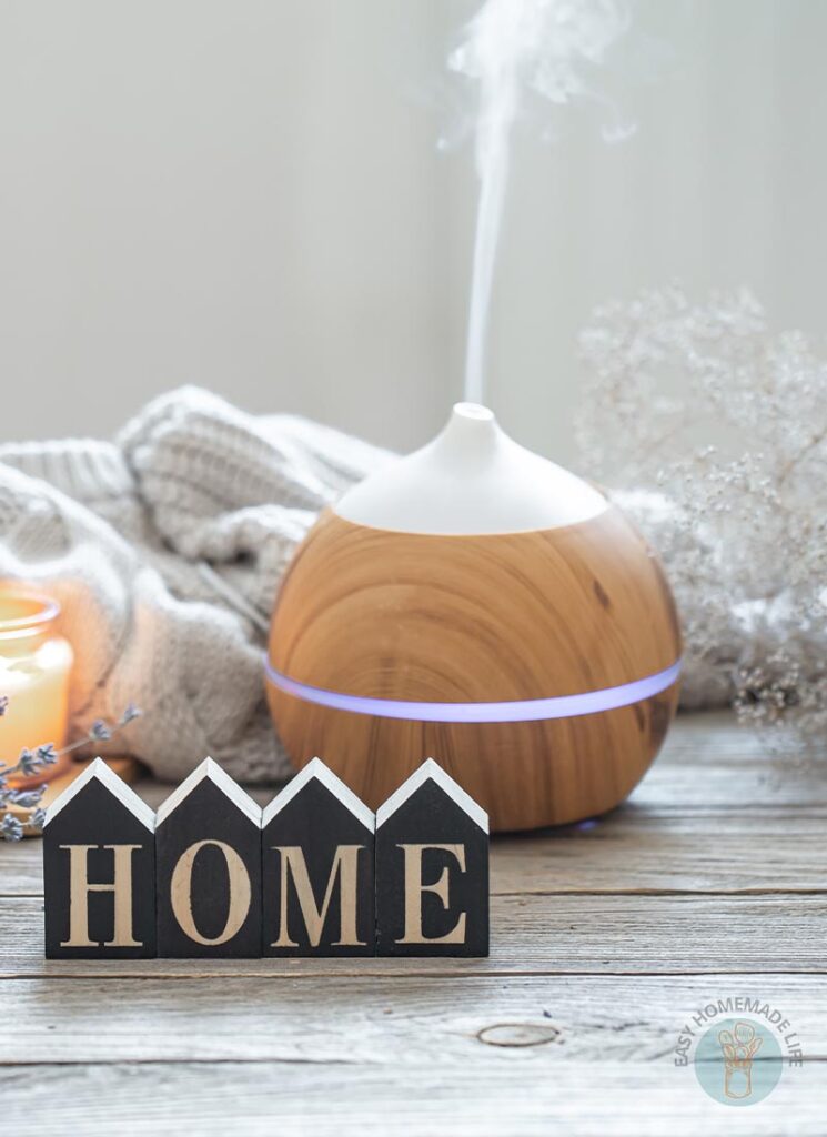 Essential oil diffuser diffusing mist next to a branch of tiny dried white flowers, a white knitted cloth and a lit candle. A "HOME" sign table decor.