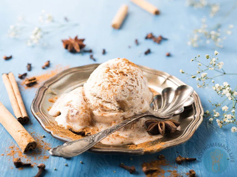 A scoop of chai ice cream in a silver plate with a spoon surrounded by cinnamon sticks, cloves and star anise on a blue table.