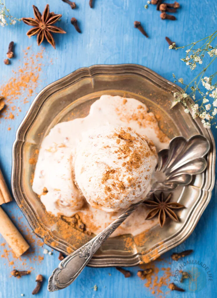 A scoop of chai ice cream garnished with cinnamon powder in a silver plate with a spoon surrounded different kinds of herbs.