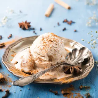 A scoop of chai ice cream garnished with cinnamon powder in a silver plate with a spoon and herbs scattered on the table.