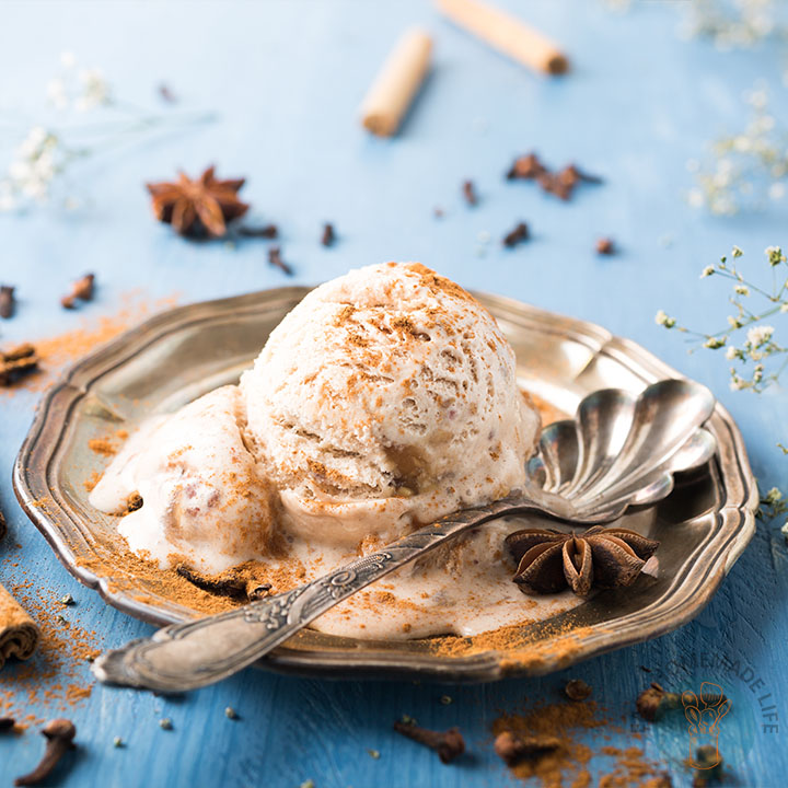 A scoop of chai ice cream garnished with cinnamon powder in a silver plate with a spoon and herbs scattered on the table.