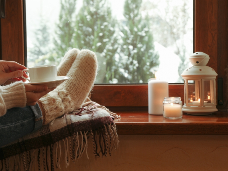 A person wearing winter socks, feet up on a pillow and holding a cup of hot drink in front of a window with lit candles and a lantern essential oil diffuser.