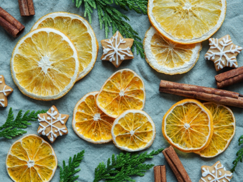 Baked orange and cinnamon with sugar cookies in snowflake shape next to pine boughs and cinnamon sticks on a blue cloth.