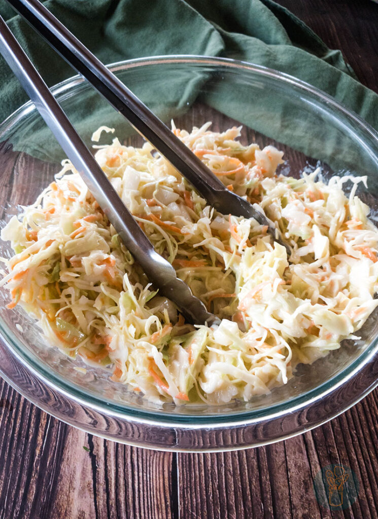Copycat popeyes coleslaw dressing in a glass bowl with metal serving tongs on a wooden table.