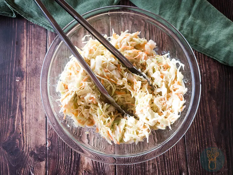 Copycat popeyes coleslaw dressing in a glass bowl with metal serving tongs next to a green cloth on a wooden table.