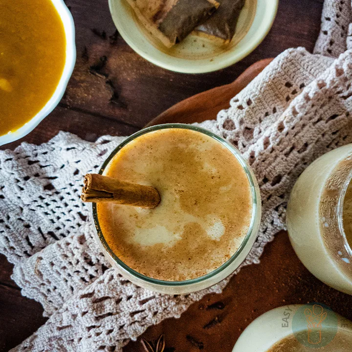 Three glasses of pumpkin chai latte with cinnamon sticks on a clay plate next to a saucer with used tea bags and a bowl with brown liquid.