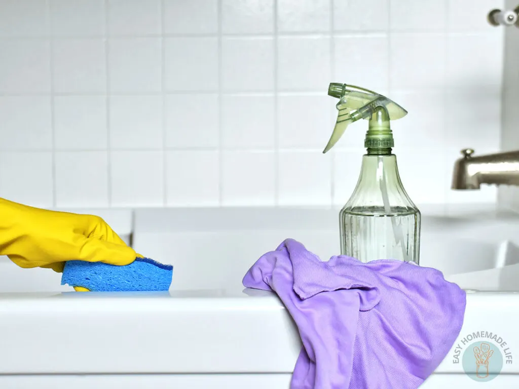 A hand wearing yellow glove holding a blue cleaning sponge appearing to clean the bath tub. Green spray bottle and purple wash cloth on the side.