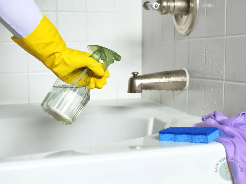 A hand wearing a yellow hand glove holding a green spray bottle. Blue sponge and purple cloth on the side.