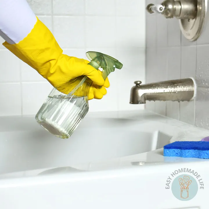 A hand wearing a yellow hand glove holding a spray bottle cleaning the bath tub.
