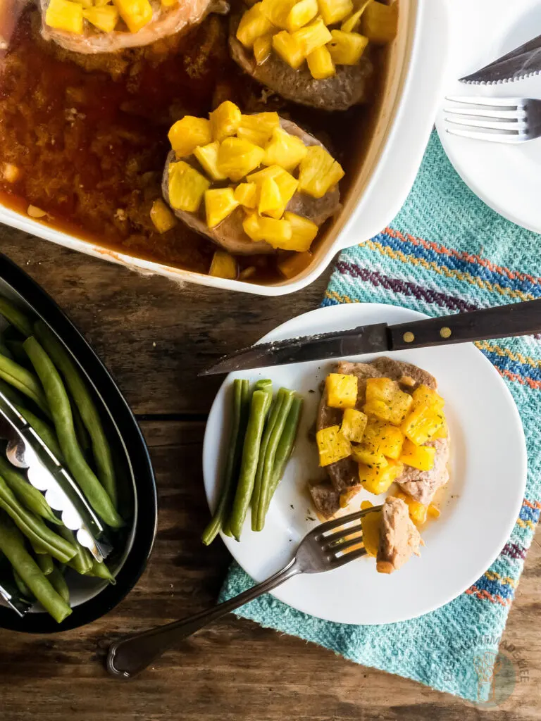 Pineapple teriyaki pork chops in a baking dish, one of them on a white plate with a fork and a knife.