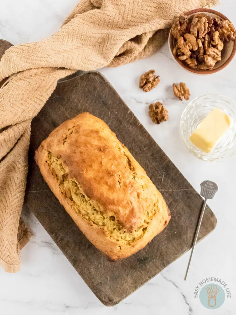 A whole loaf of banana bread on a wooden chopping board next to a slice of butter and a bowl of walnuts.