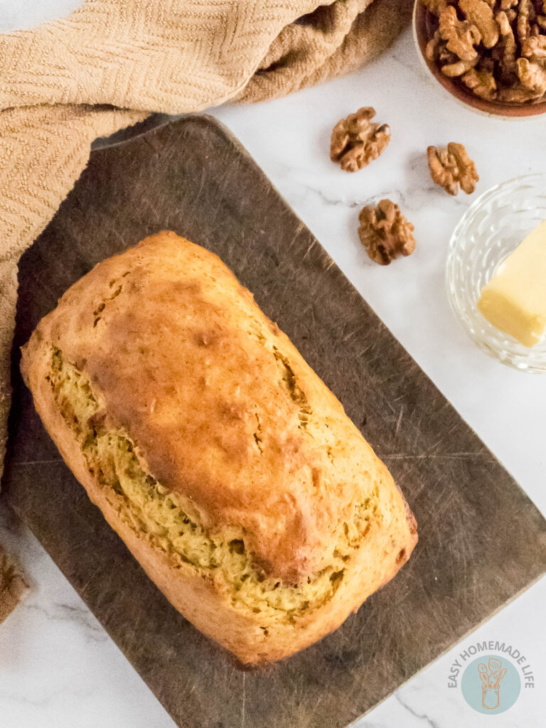 A whole loaf of banana bread on a wooden chopping board next to a slice of butter and a bowl of walnuts.