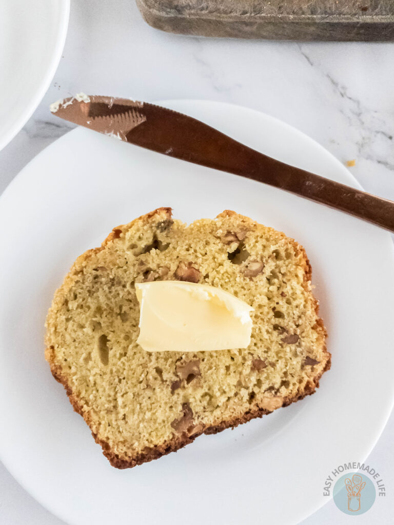 A white plate with a slice of banana bread without baking soda with a butter topping on a white plate with a bread knife next to it.