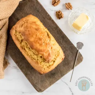 A whole loaf of banana bread on a wooden chopping board next to a slice of butter and pieces of roasted walnuts.