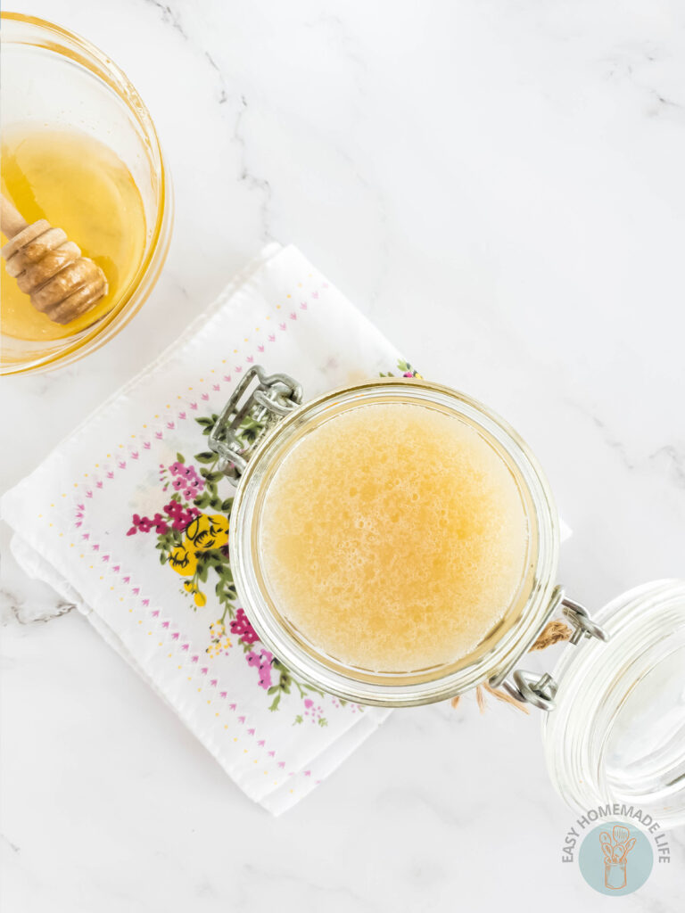 An open jar of honey body scrub next to a glass bowl with honey and honey dipper.