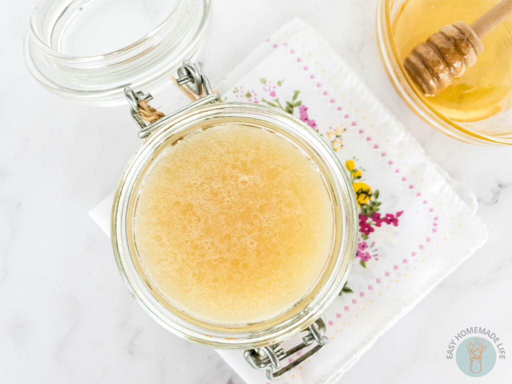 An opened jar of honey body scrub next to a bowl of honey with dipper.