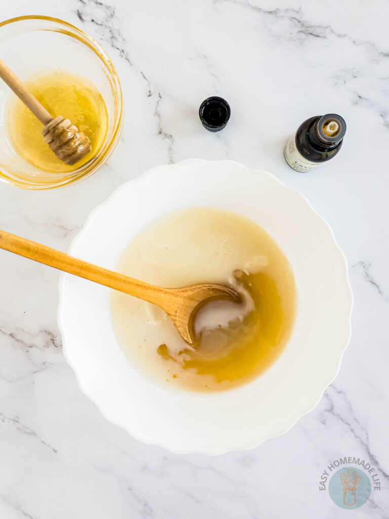 A white bowl with a wooden spoon mixing a white mixture of honey body scrub.