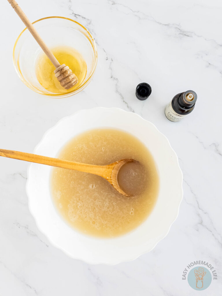 A white bowl with a wooden spoon mixing a white mixture of honey body scrub next to a bottle of essential oil.
