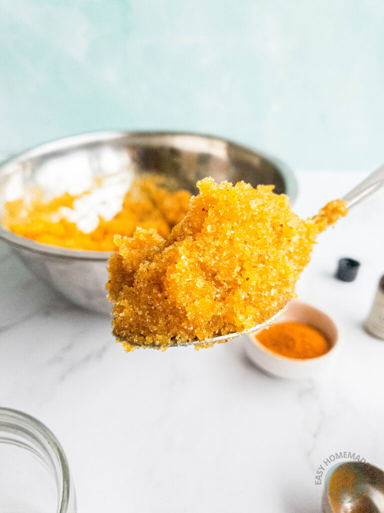 A spoonful of turmeric body scrub. A stainless steel mixing bowl and ingredients at the background.