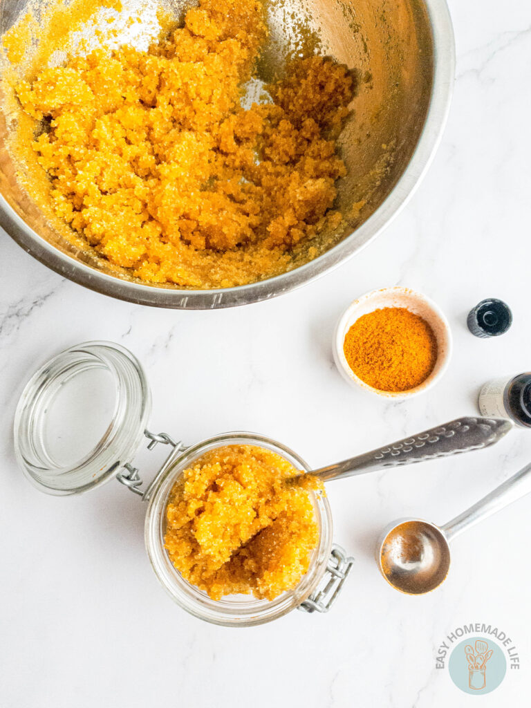 A jar of turmeric body scrub next to a stainless steel mixing bowl, turmeric powder and measuring spoon.