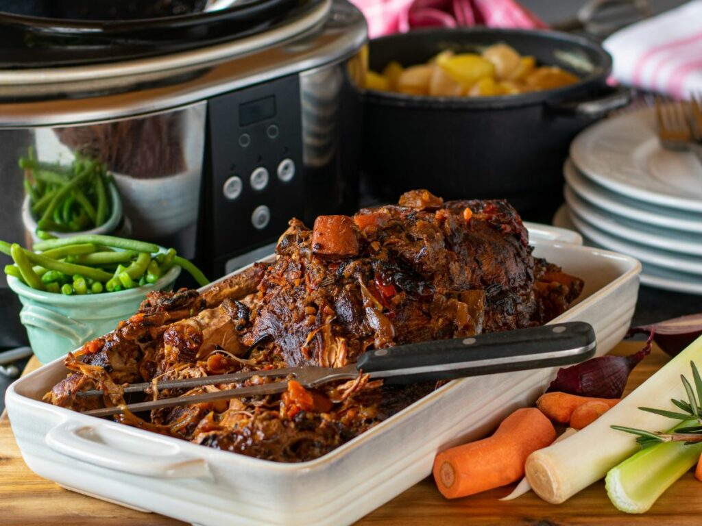 Pot roast dish next to a variety of vegetables with a slow cooker in the background.