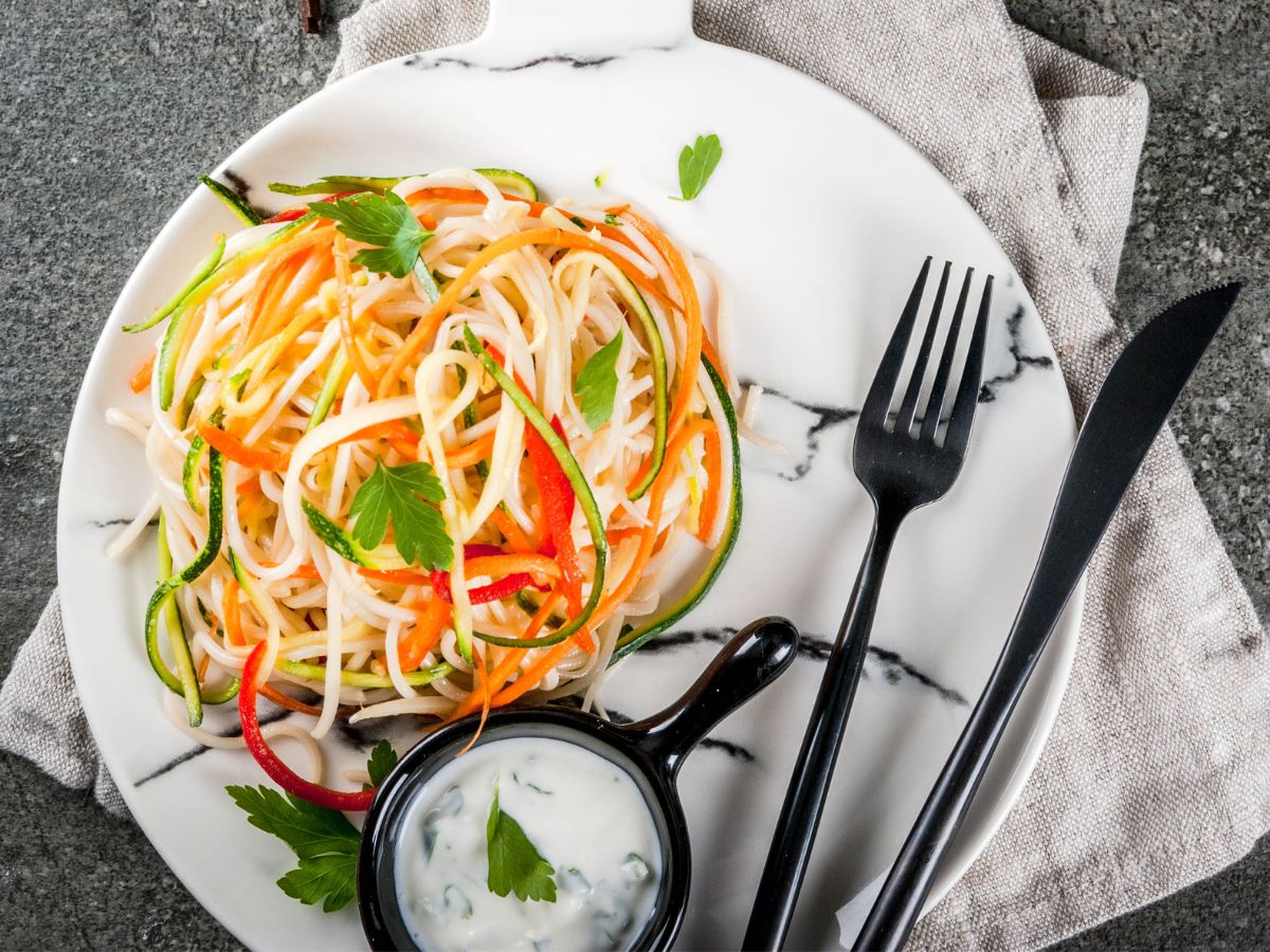 Spiralized veggie noodles with dipping sauce on a plate with knife and fork.