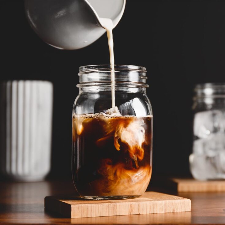 Milk in white pitcher being poured into black iced coffee.