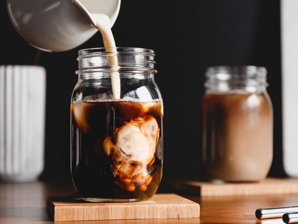 Milk in white pitcher being poured into black iced coffee in jar on wooden block coaster.