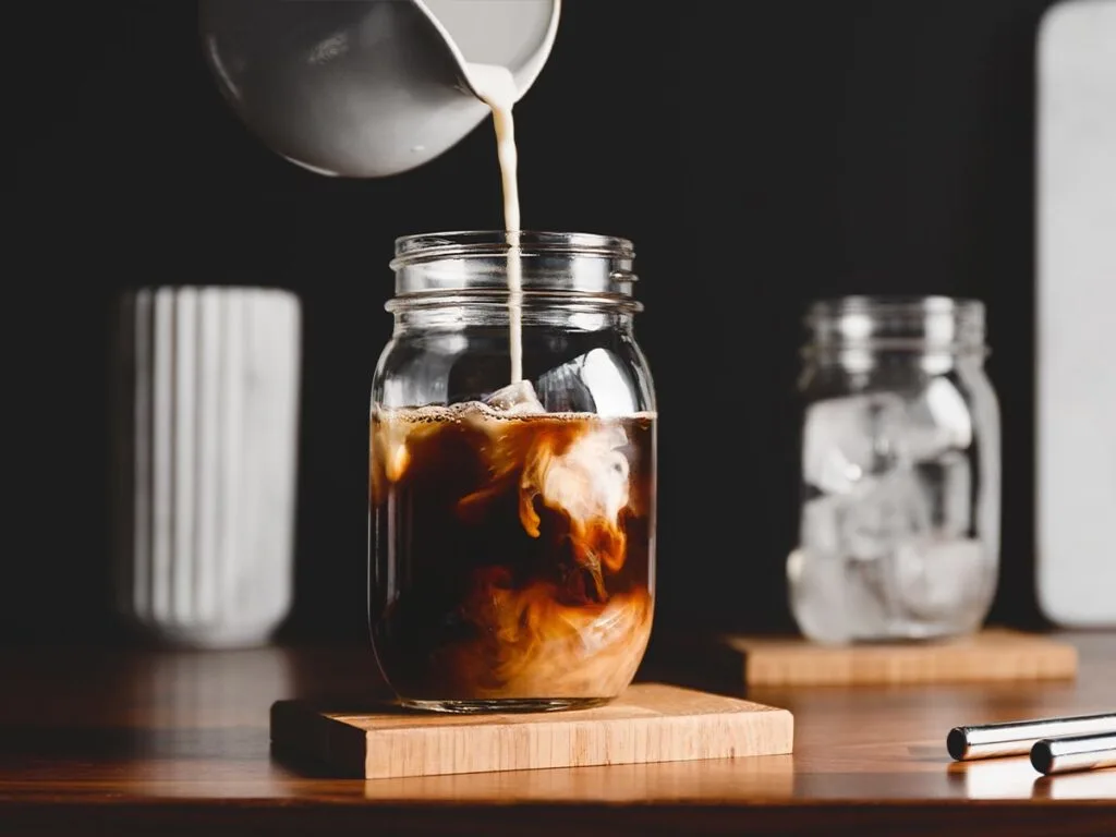 Milk in white pitcher being poured into black iced coffee in jar on wooden block coaster.