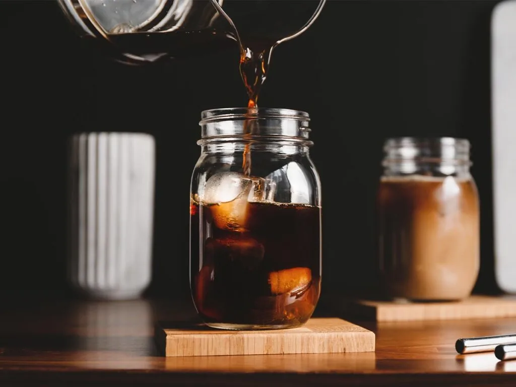 Cold brew coffee being poured over ice cubes in a glass on wooden block coaster.