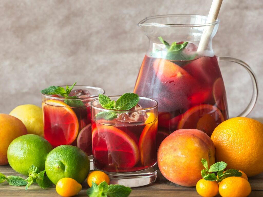 A pitcher of homemade sangria next to two glasses full of the same drink surrounded with fresh ingredients.