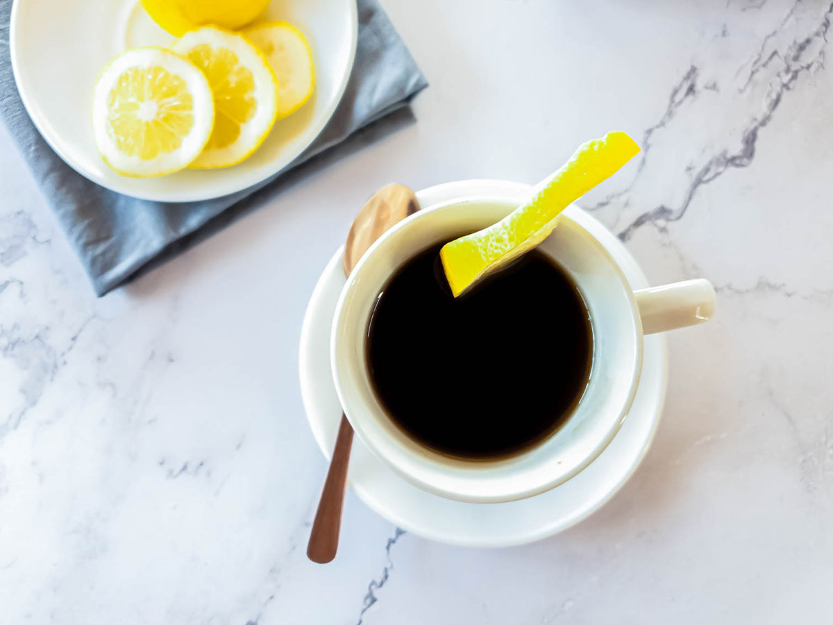 Lemon slices on plate with grey napkin, fresh lemon, and espresso in white cup.