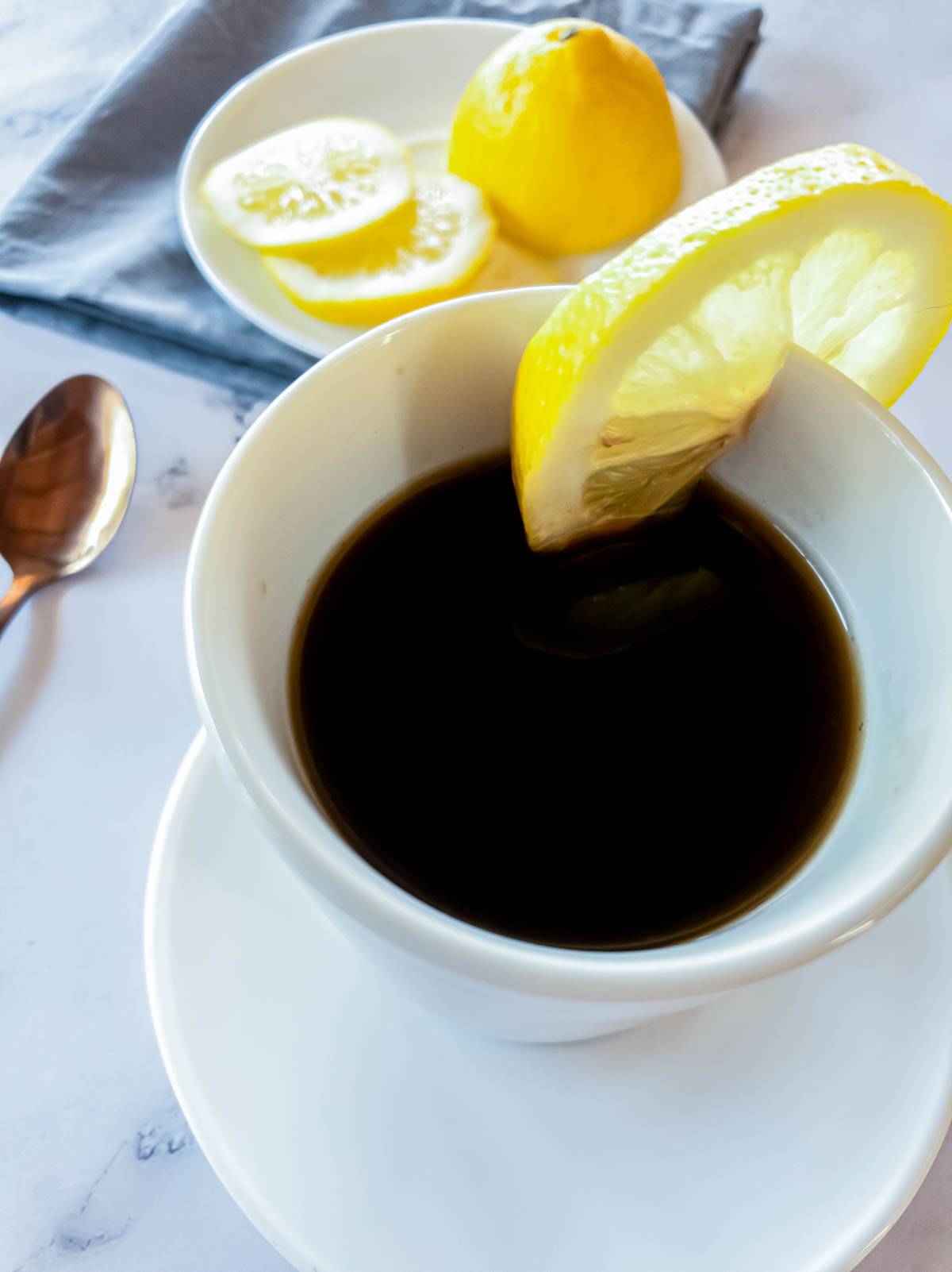 Hot cafe Romano in espresso cup with white saucer, sliced lemon in background.