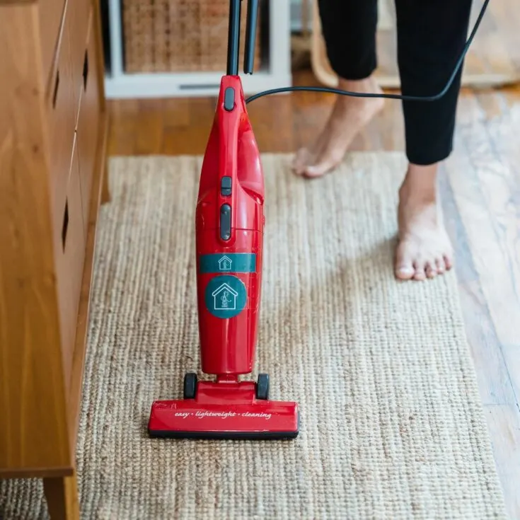 Barefoot woman on tan carpet vacuuming with red vacuum.