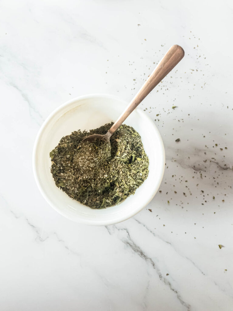 Dry herb mixture with copper spoon in white bowl.