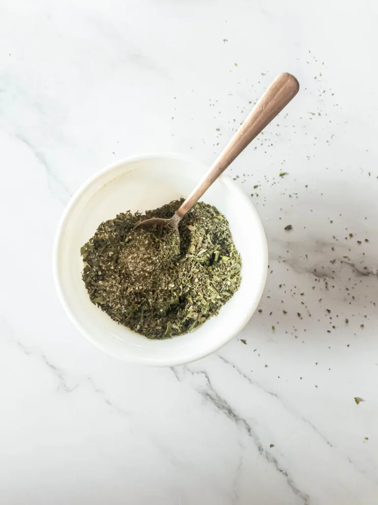 Dry herb mixture with copper spoon in white bowl.