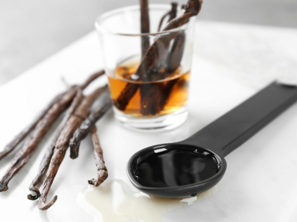 Vanilla extra in measuring spoon with vanilla beans on countertop.