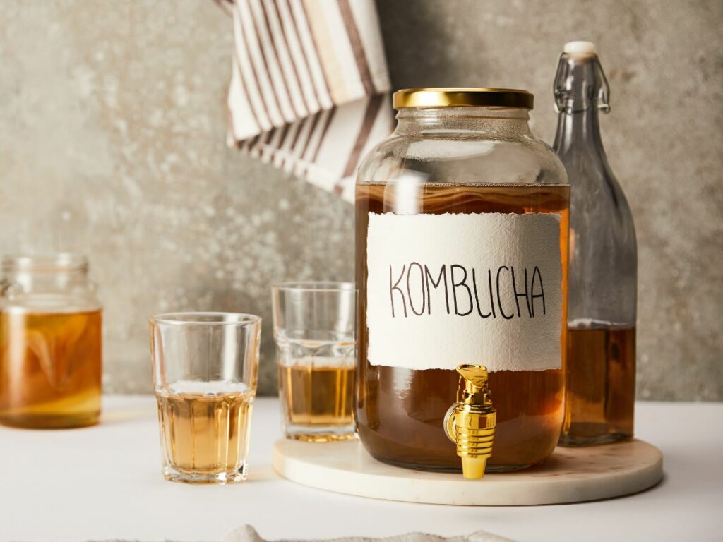 A glass dispenser of kombucha with golden faucet next to a glass bottle with brown liquid and two half filled glasses with the same liquid drink.