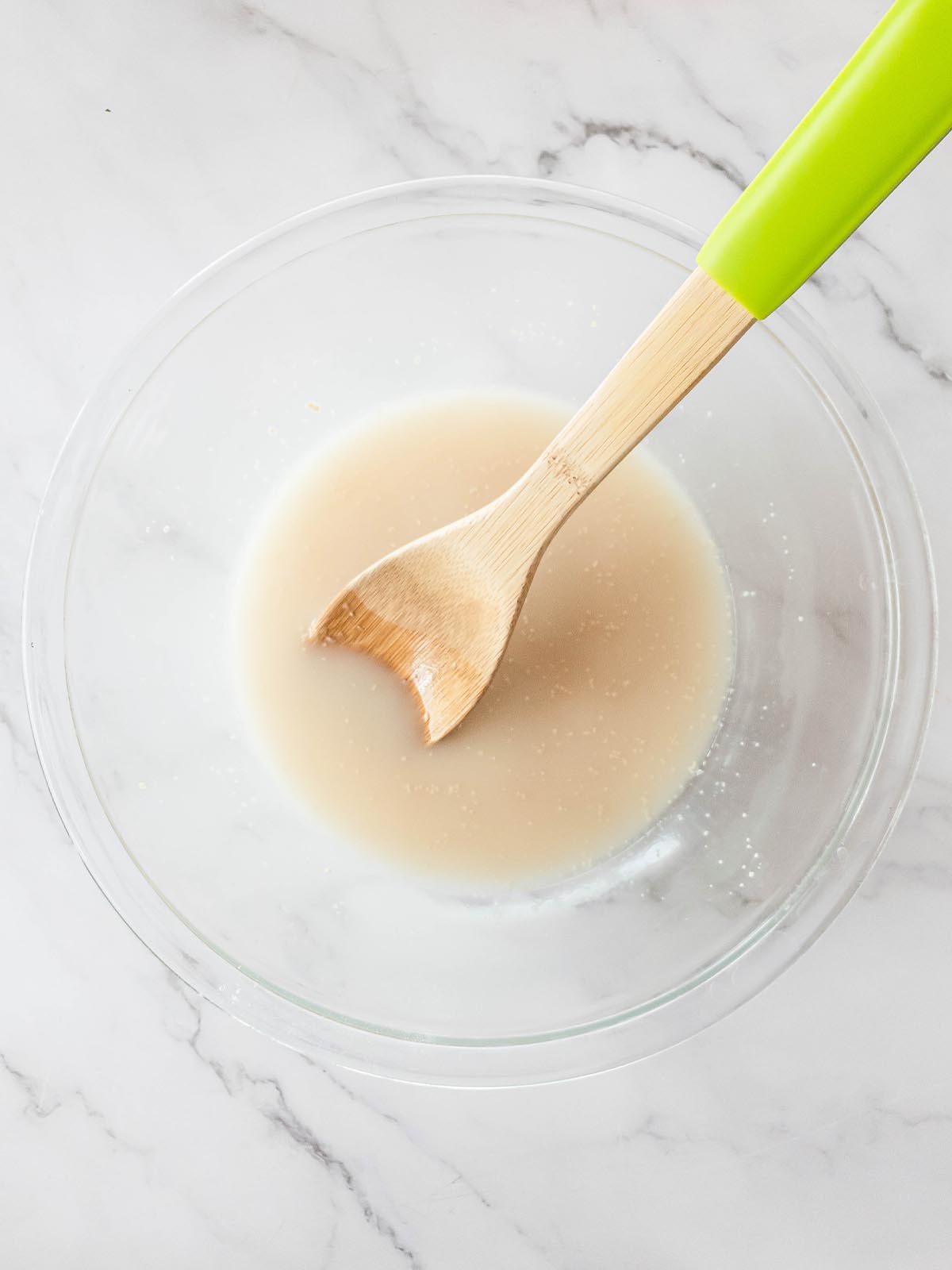 A green wooden spoon stirring white mixture in a transparent bowl.