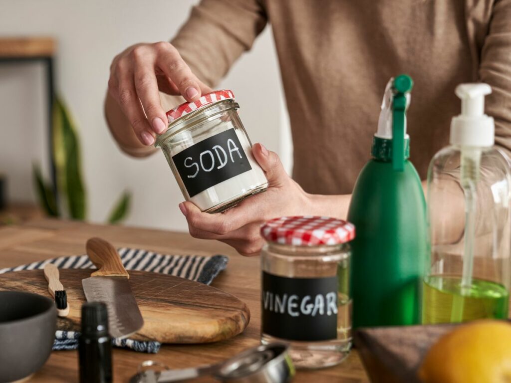 A woman is holding a jar of baking soda on a kitchen table with natural cleaning supplies.