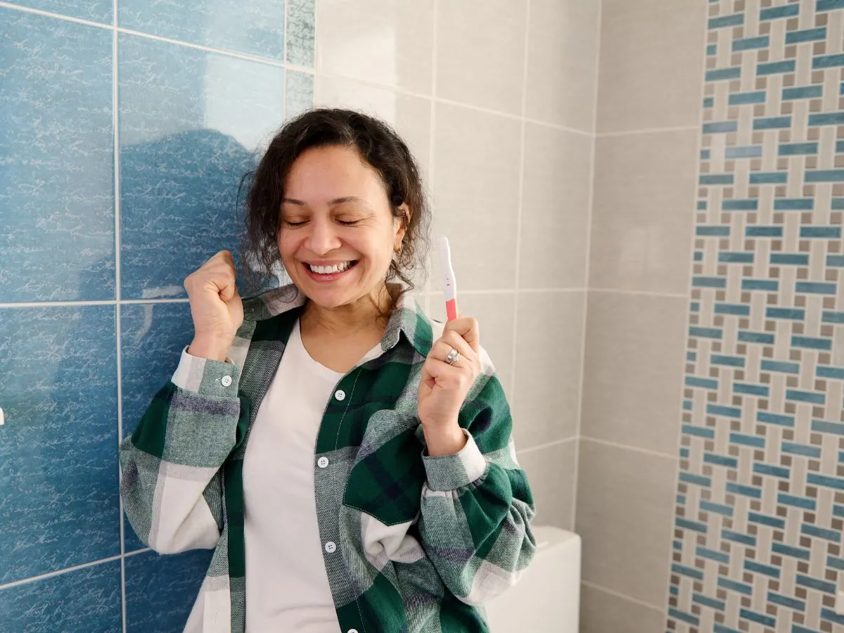 A happy woman holding a pregnancy test stick in a bathroom.