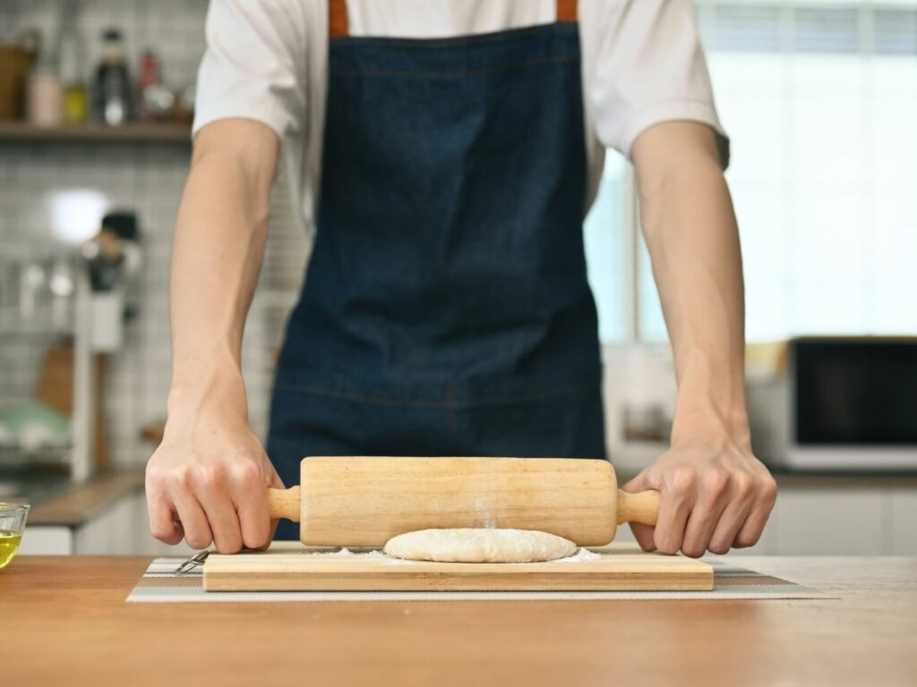 A man in an apron is rolling dough on a wooden board using an alternative to a rolling pin.